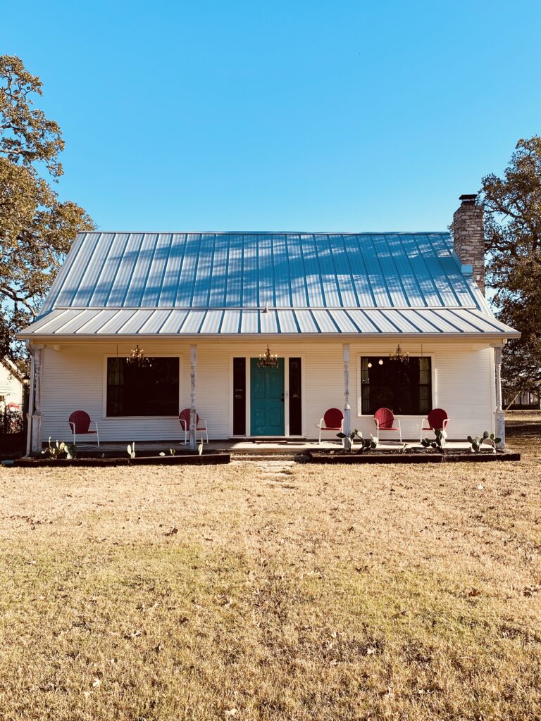 Small white house with tin roof. Two windows flank a teal front door. Four white columns on the porch. Also on the porch are 5 red retro looking porch chairs. 