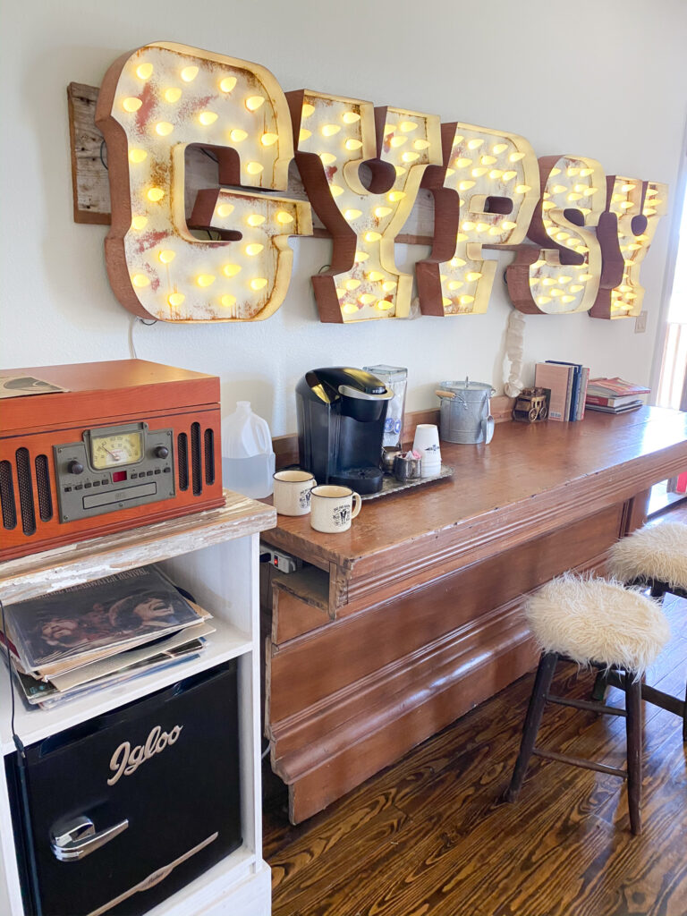 Large sign that reads "Gypsy" hangs above a wood coffee bar. Two stools in front with white furry seats. To the left of the coffee bar is a record player. 