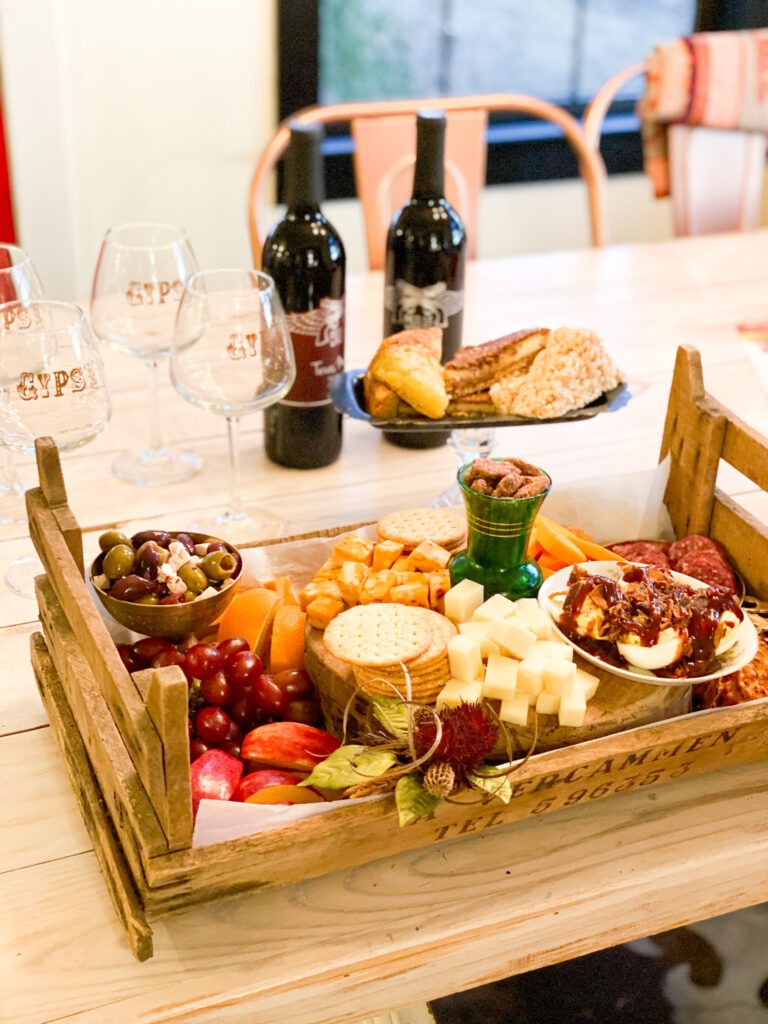 Close up of table with wine glasses in the upper left. Next to the wine glasses are two bottles of wine.  In front of the glass and wine is a wood crate filled with various fruits, cheeses, and meats. 