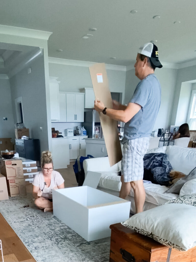 man on the right of the picture is holding a piece of cardboard standing in a living room. on the floor to his left is a woman sitting on a floor holding a hammer assembly one of the closet systems. 