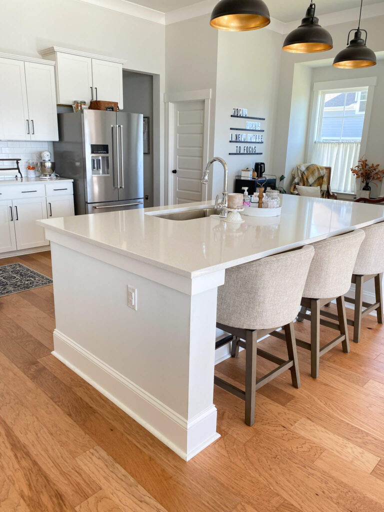white kitchen island with three gray bar stools. there are three black pendant lights hanging above. a stainless steel fridge sits in front of on the left side of photo next to white cabinets. the floor is a sandy oak color.
