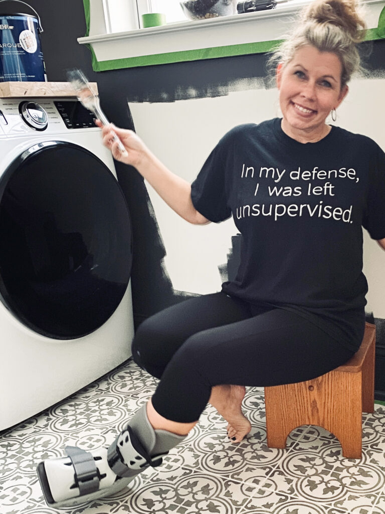 author wearing a black t shirt that reads "in my defence I was left unsupervised" sitting on a stool wearing a plastic cast boot on left foot holding a paint brush in her right hand sitting next to a dryer in front of a partially painted wall. 