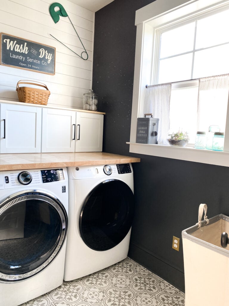 Laundry room, on the left side of the phot are a set of white washer and dryer. There is a blonde colored counter on top of the wsher and dryer and on top of that is a small set of cabinets with a shelf. On top of the shelf is a woven basket in the middle and to the right, a jar of gray dryer balls. In the middle of the picture is a white window. The walls are painted Iron Ore a dark gray black color. On the bottom right of the photo is a industrial looking canvas laundry hamper on wheels. the floor is Tile with a gray and white design.