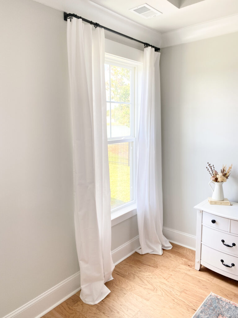 White curtains hanging from black curtain rods on the left. White bedside table with black knobs to the right. On top of the white side table is a white pitcher filled with fall florals on top of a cream colored book. Flooring is a lighter colored wood. 