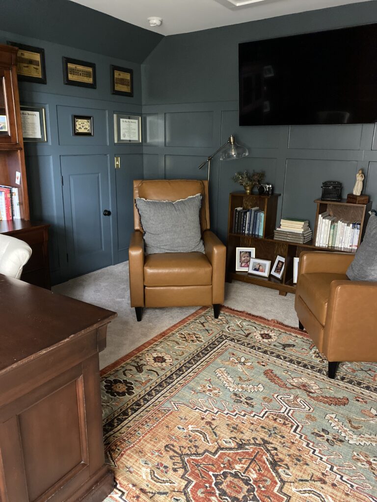 Angle of the room showing both cognac colored chairs, book shelf with books and pictures. A brass floor lamp with a glass shade is behind the chair. Hanging above the book case on the right of the photo is a TV mounted on the wall. On the left of the chair hanging on the wall are several small pictures. 