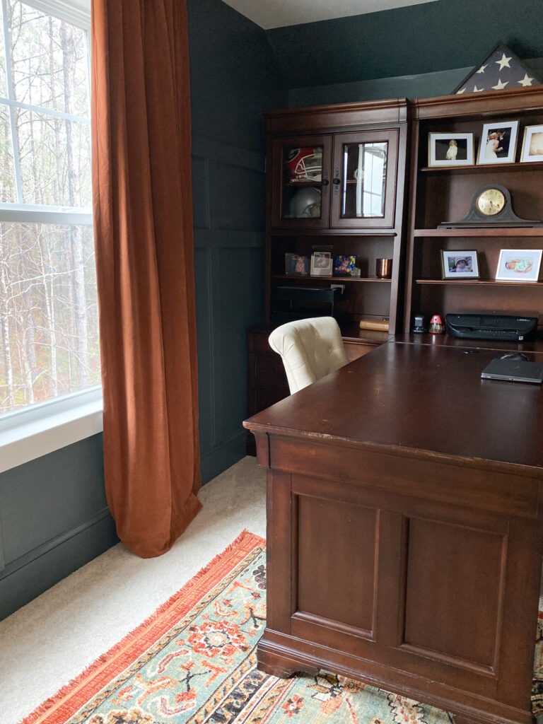 Brown desk on the right side of the photo with a light colored chair pulled in. There are several shelves above the desk that are full of mementos. Behind the chair on the left are copper colored velvet curtains hanging by the window. Beyond the window are woods. Beneath the desk is a jewel colored rug that matches the curtains, desk, and wall color. 