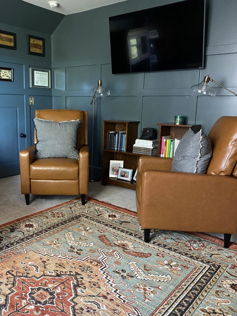 BEHR by Brooklyn painted on the walls behind two cognac colored brown chairs. Under the chairs is a rug with oranges, teal, browns, and white. 