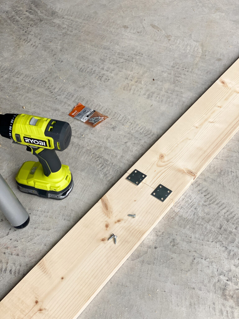 Two pieces of wood laying on a concrete floor with two metal brackets laying on top to conjoin them together. A drill is sitting next to the two boards. 