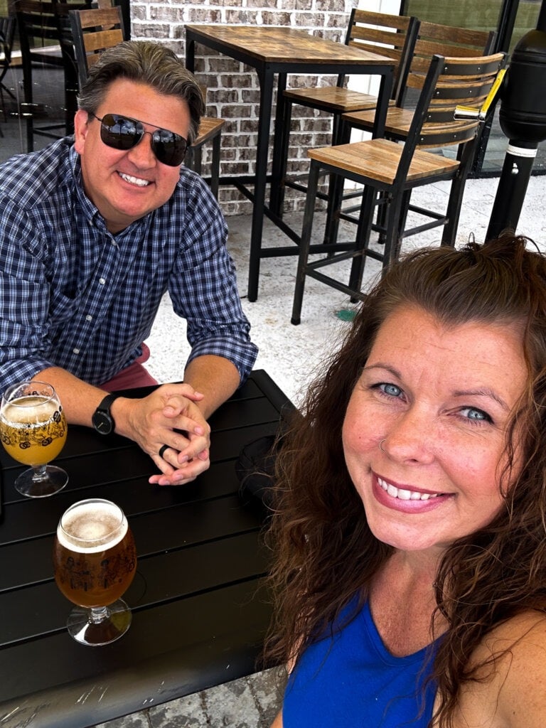 My husband and I sitting at a table outside of a brick building with two glasses of beer between us. 